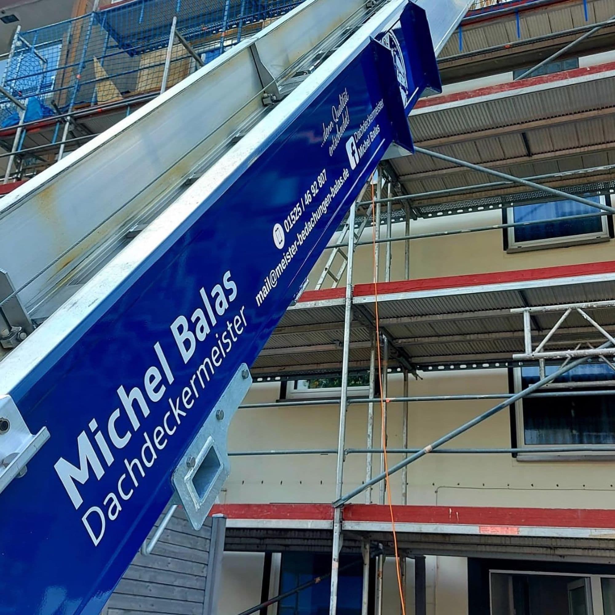 Blauer Schrägaufzug mit Beschriftung eines Dachdeckermeisters vor einem eingerüsteten Haus