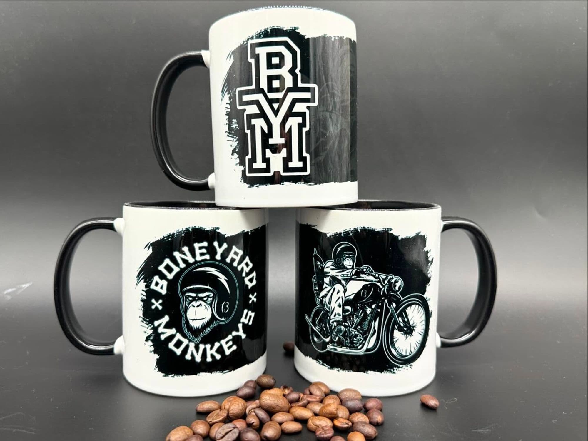 Drei weiße Tassen mit schwarzem Henkel, bedruckt mit Motiven von Boneyard Monkeys, davor Kaffeebohnen.