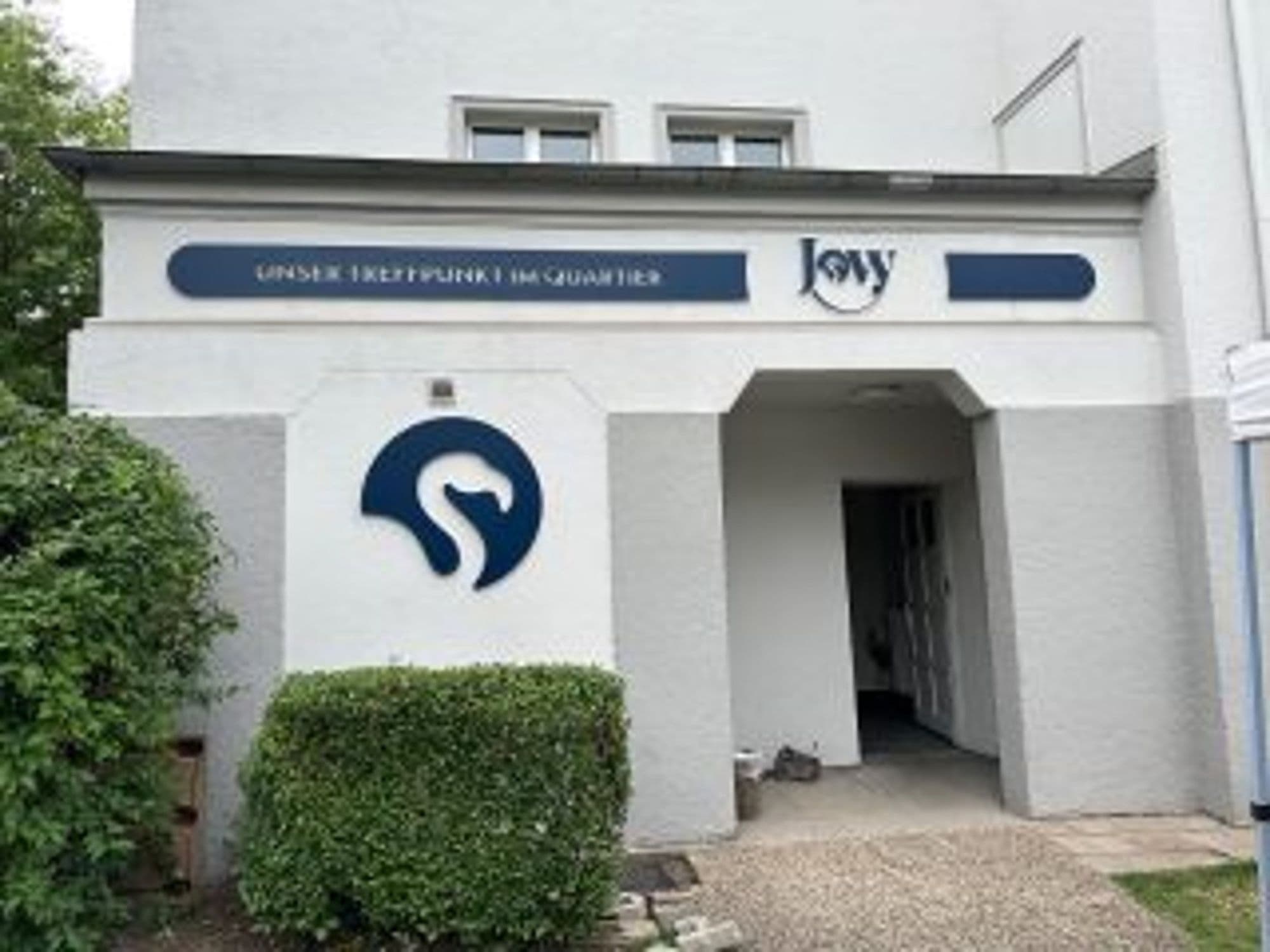 Eingangsbereich des Jovy Quartierstreffs mit gefrästem Logo, Schriftzug und Schriftband in Dunkelblau an der Fassade.