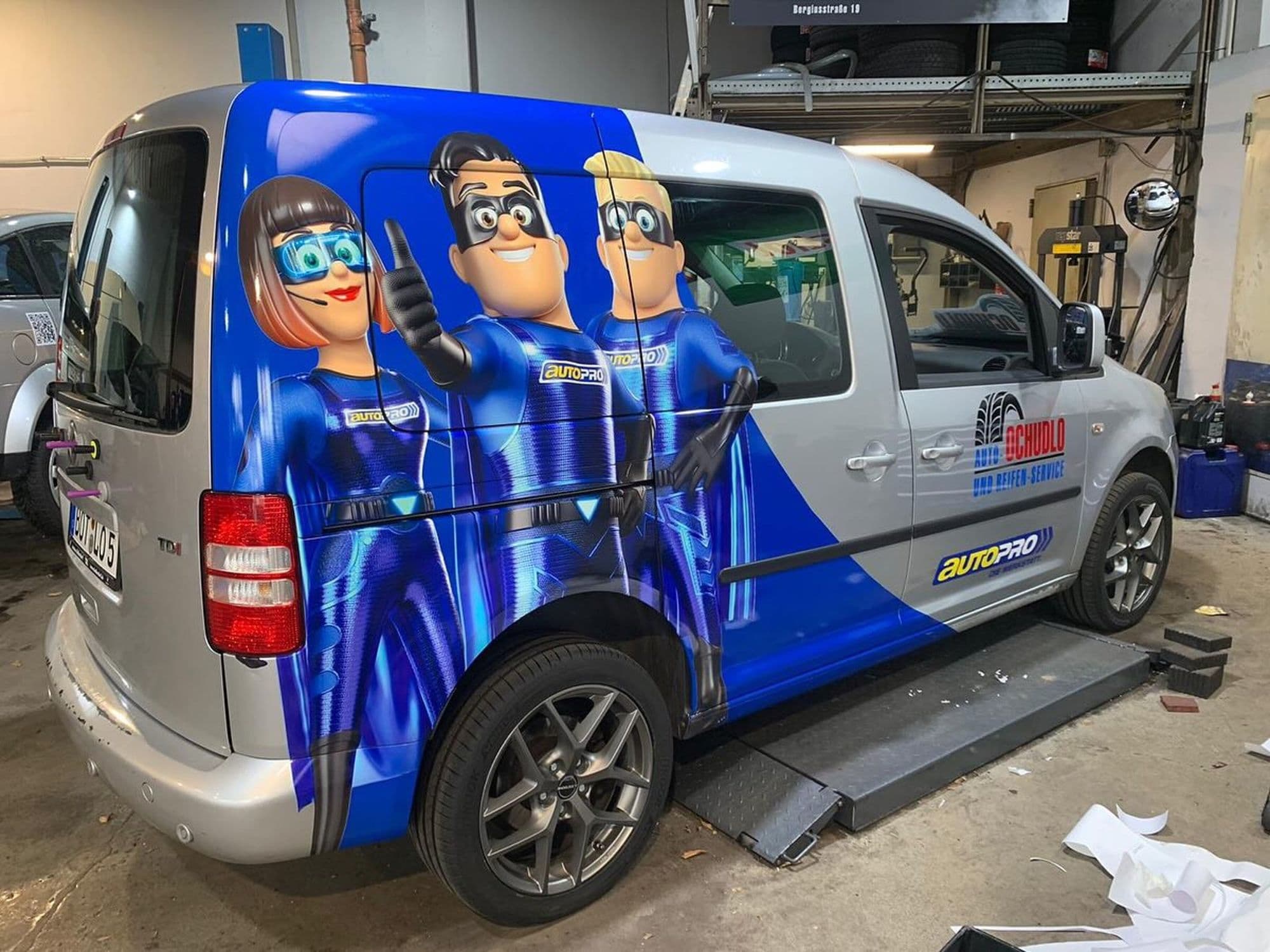 Grauer Transporter mit Comicfiguren in Superheldenkostümen in einer Werkstatthalle