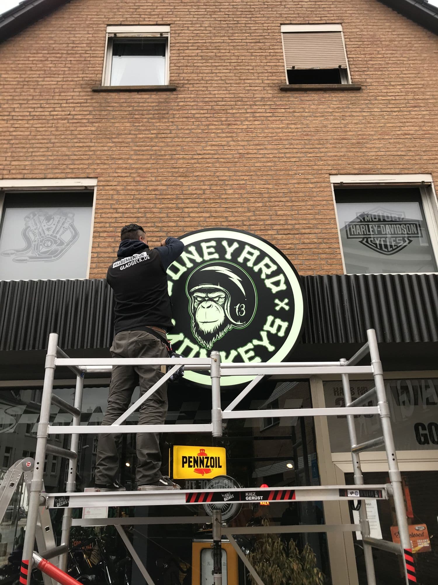 Monteur auf einem Gerüst bringt einen runden, grün leuchtenden Leuchtkasten von Boneyard Monkeys an einer Klinkerfassade an.