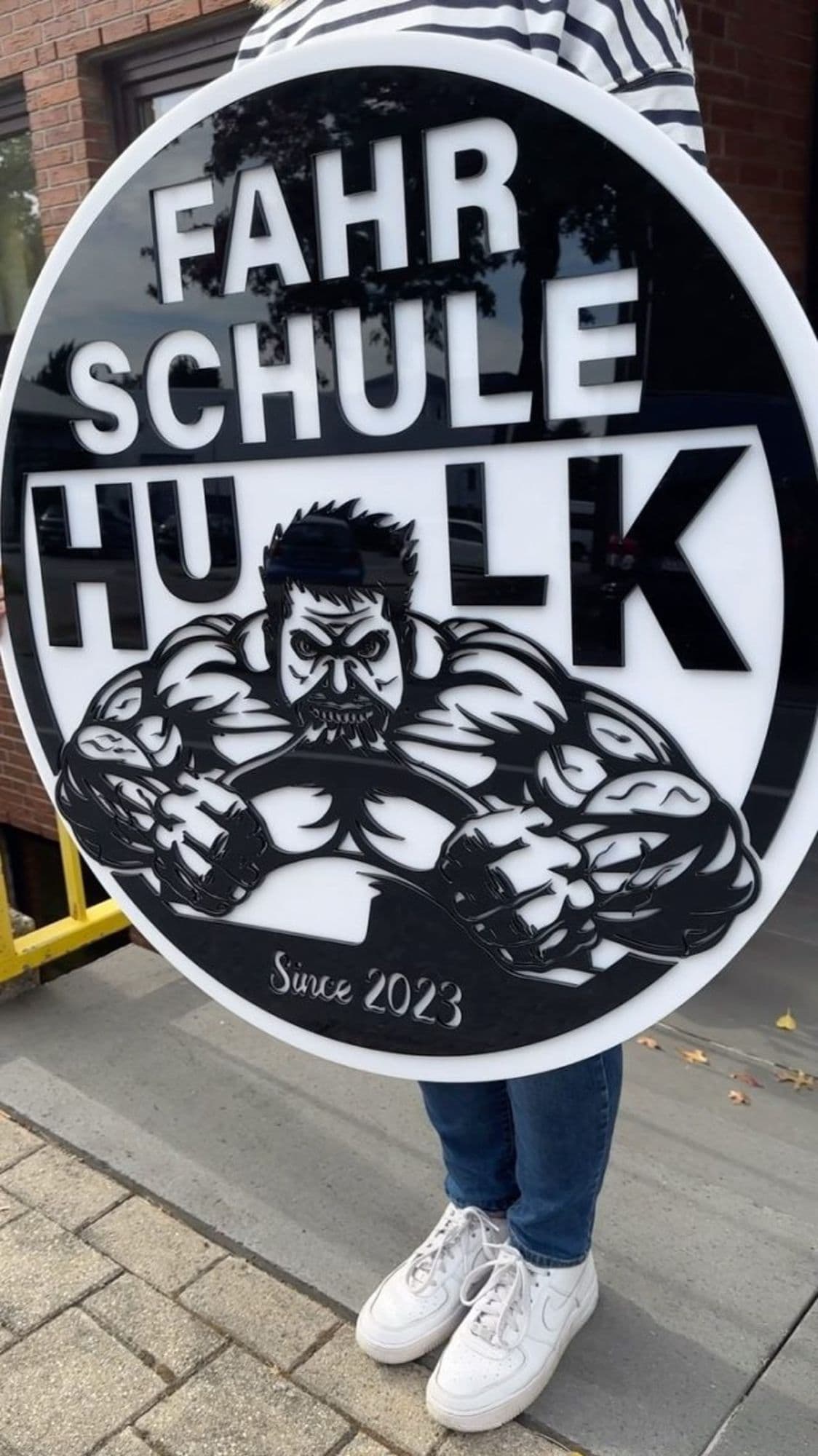 Rundes, mehrschichtig gefrästes Logoschild der Fahrschule Hulk mit erhabenen Buchstaben in Schwarz und Weiß.