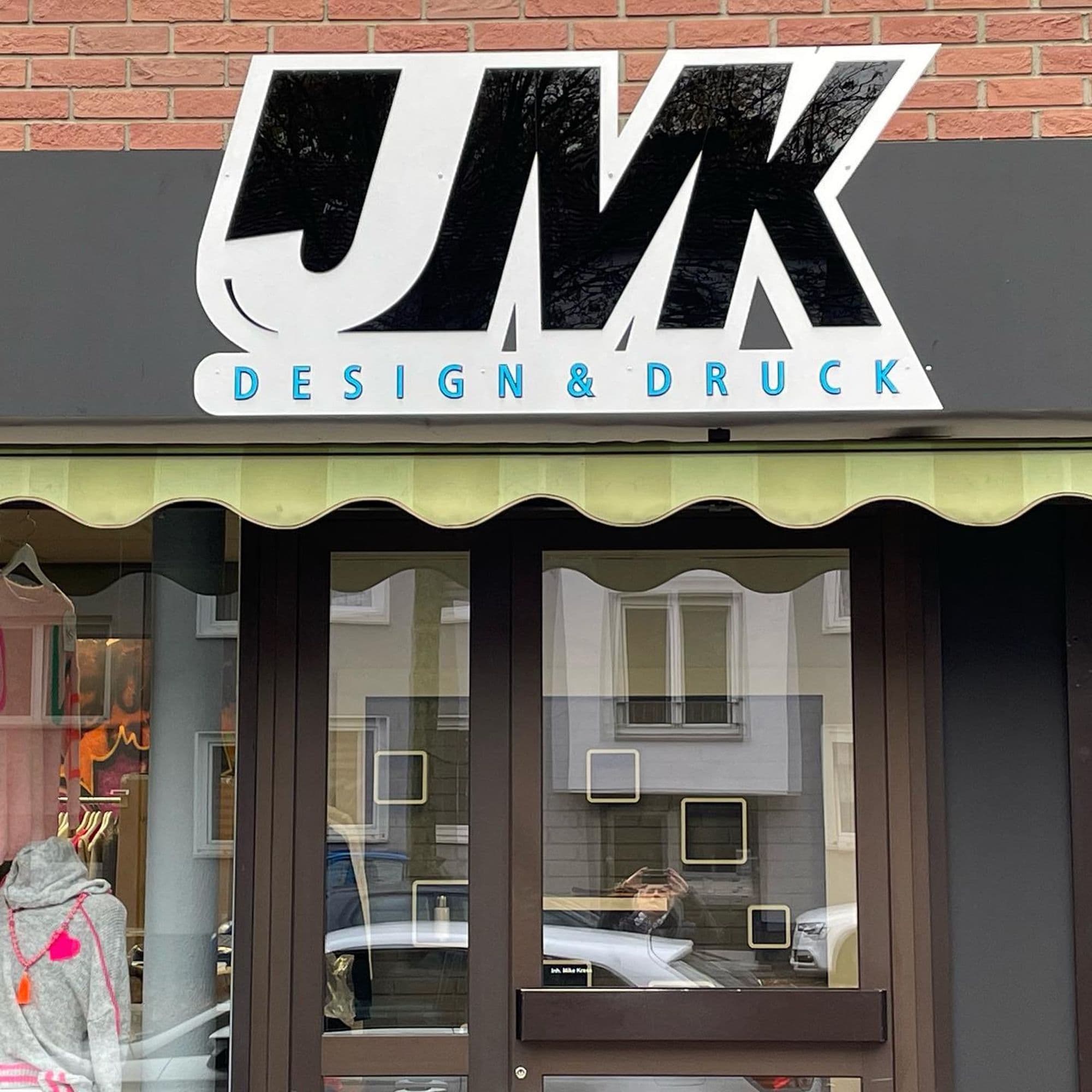 Ladenlokal von JMK Design & Druck mit großem Logoschild über Schaufenster und Eingangstür.