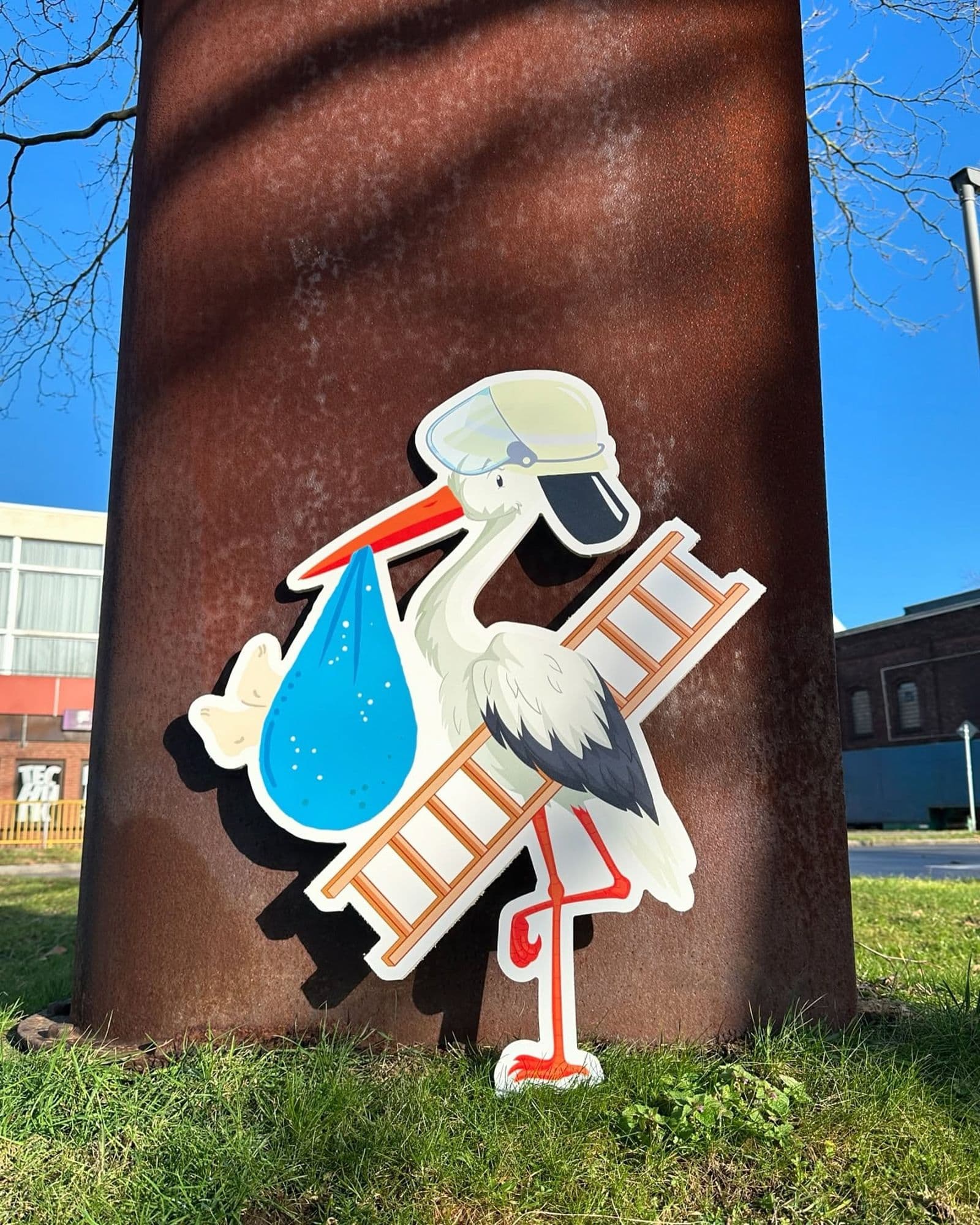 Konturgeschnittener Aufsteller in Storchform mit Feuerwehrhelm, Leiter und blauem Bündel, aufgestellt im Freien.