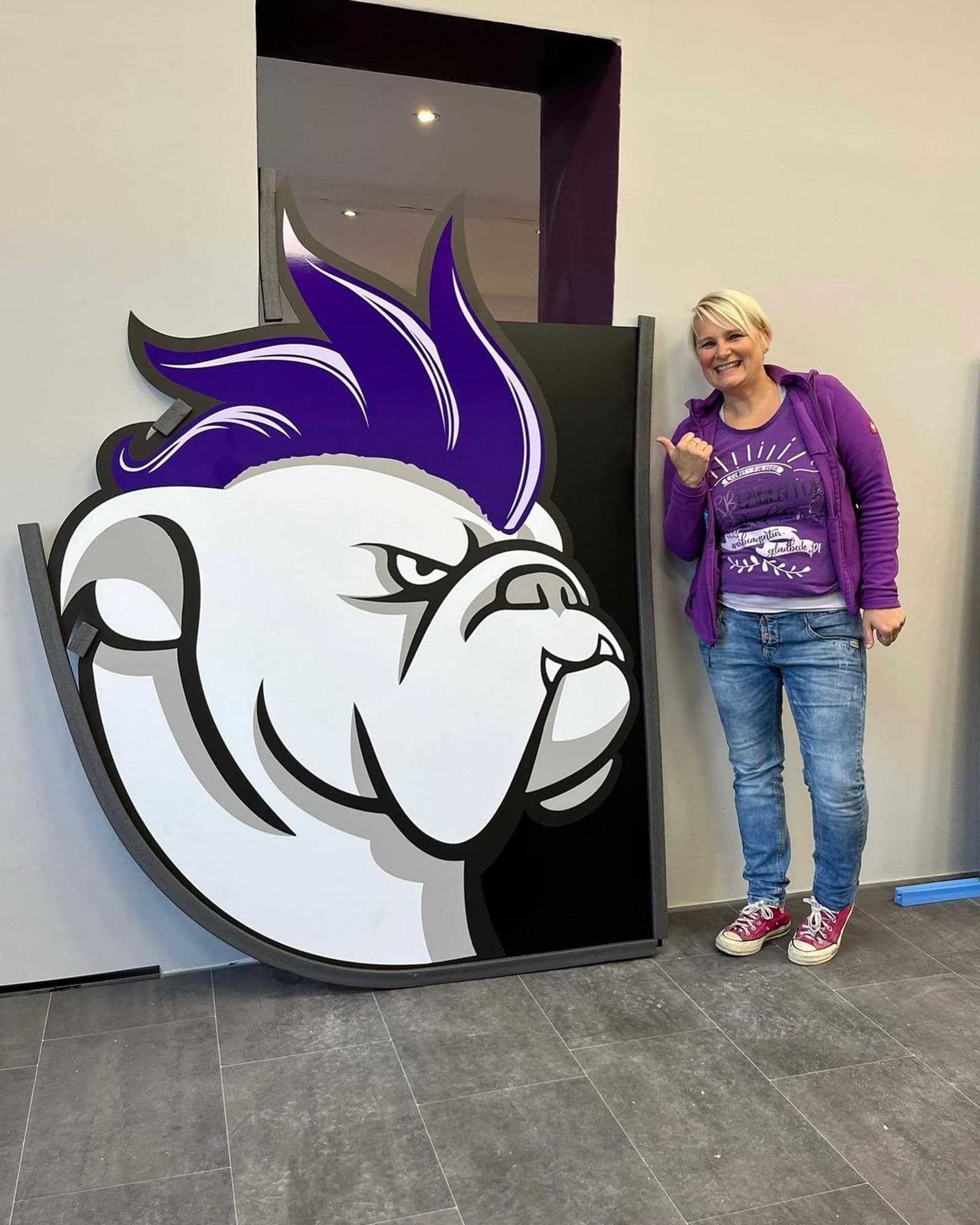 Frau mit blondem Kurzhaarschnitt und lila Kleidung steht neben einem großen Bulldoggen-Logo mit lila Haaren.