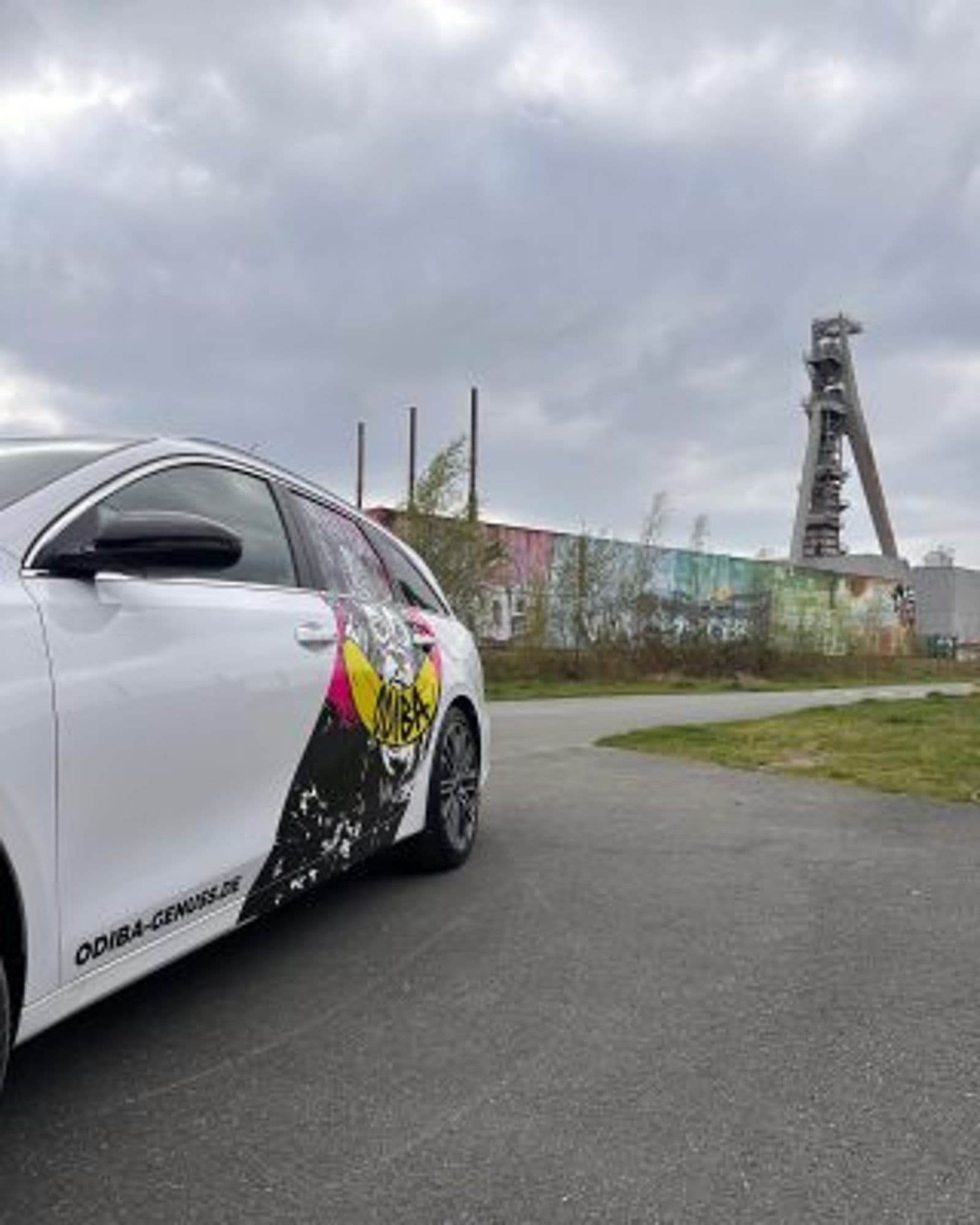 Weißes Auto mit farbiger Teilbeklebung, im Hintergrund ein Förderturm