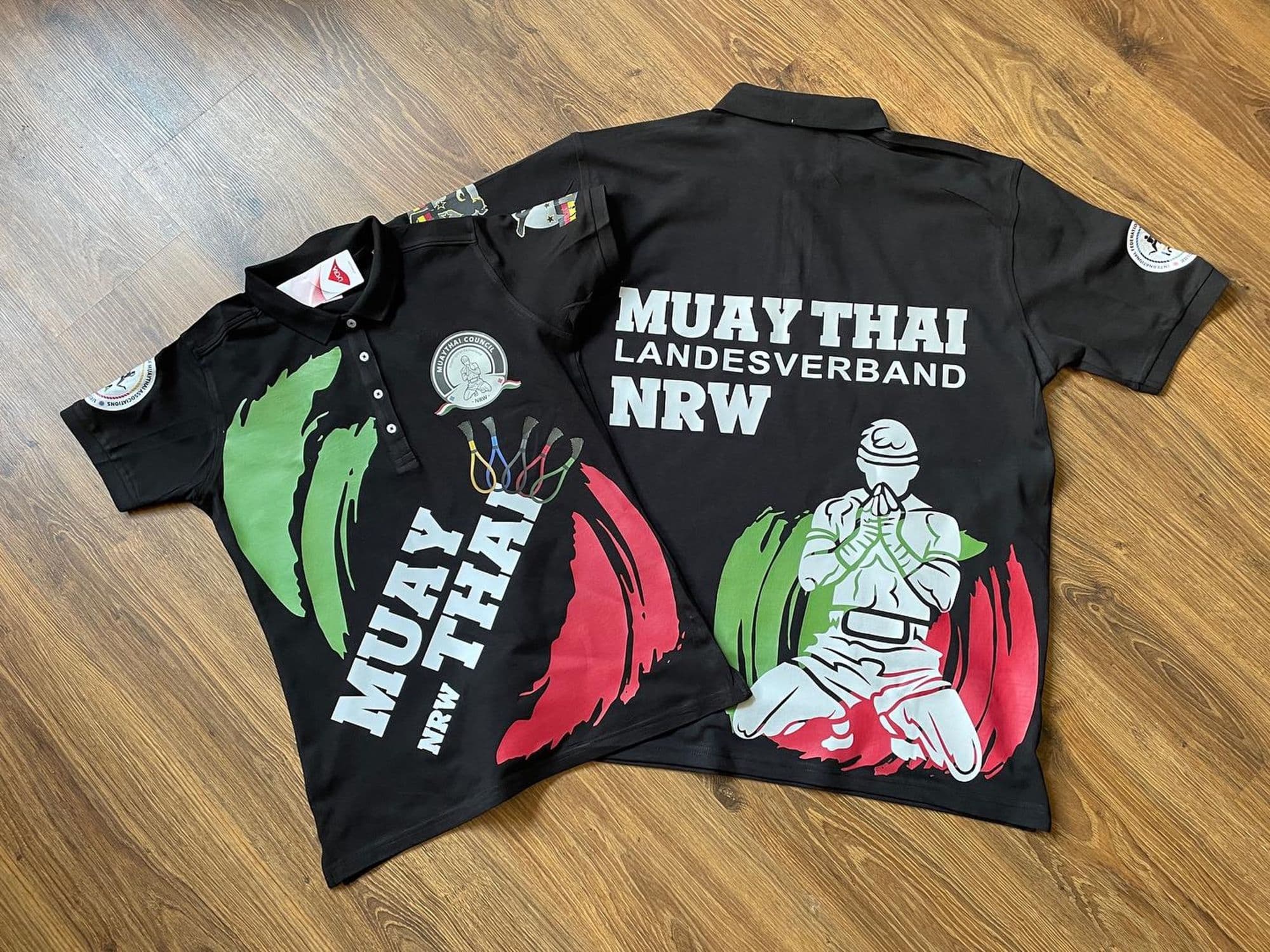 Zwei schwarze Poloshirts des Muay Thai Landesverbands NRW mit großflächigem Druck in Grün, Rot und Weiß auf Holzboden.