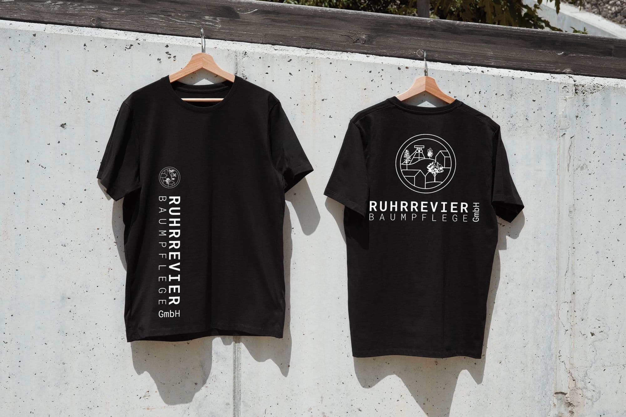 Zwei schwarze T-Shirts mit bedrucktem Firmenlogo auf Bügeln an einer Betonwand